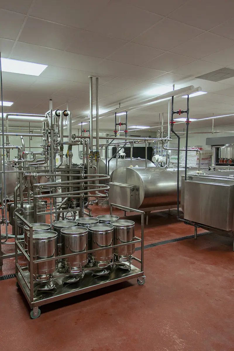 A dairy processing hall with stainless steel milk tanks, pipework and a cart of steel milk cans on a red floor