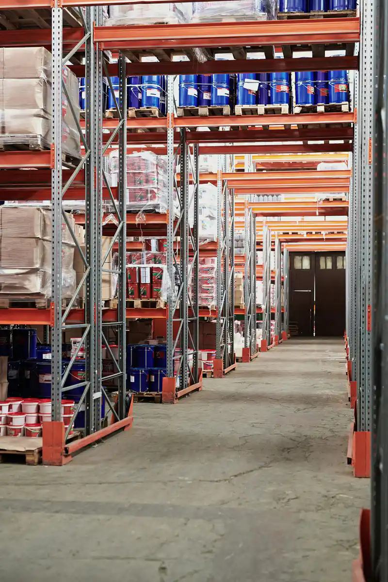 A warehouse aisle between tall steel racking, with pallets and drums on the shelves